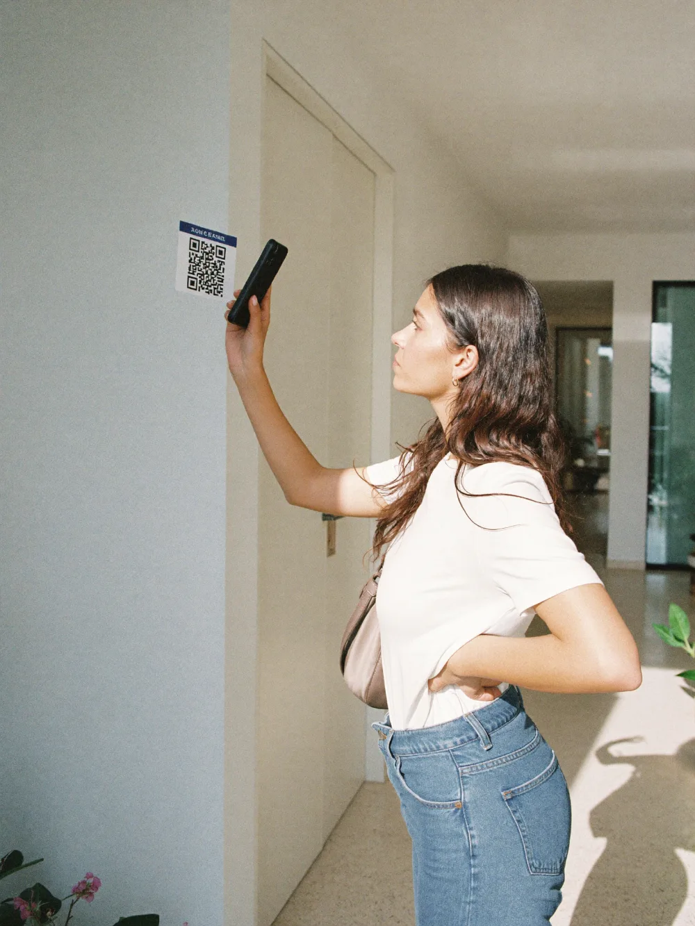 A parent scanning a printed QR code on the wall with her phone camera in a sunlit hallway